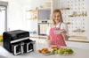 A woman in a kitchen is looking at her phone while holding a mug. An air fryer and food are on the counter. Podgląd zdjęcia produktu