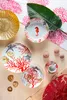 A collection of coral-themed tableware, including plates, a teacup and saucer, and decorative shells, arranged on a light beige surface with a coral-patterned fabric in the background. Voir les images du produit