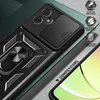 A close-up view of a black smartphone case with a ring stand and camera cover, placed on a textured surface with a green background. Produktbild-Vorschau