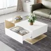 A modern living room scene with a white high-gloss coffee table with wood accents and a drawer pulled out, containing headphones and a book. A bouquet of white flowers sits on top of the table, and a green leather sofa is visible in the background. Voir les images du produit