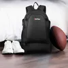 A black backpack with a shoe compartment, a pair of white sneakers, and a football are arranged on a wooden floor next to a white bed. Produktbild-Vorschau
