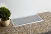 A silver grate cover is placed on a pebble-covered surface next to a white wall and a potted plant. Produktbild-Vorschau