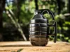 A black mug shaped like a hand grenade sits on a wooden surface in a forest setting. Voir les images du produit