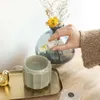 A hand is adding perfume oil to a ceramic diffuser, with a vase of dried flowers in the background. Produktbild-Vorschau