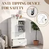 A white cabinet with doors and drawers, featuring an anti-tipping device for safety. The cabinet is shown in a room setting with a couch and a plant. Produktbild-Vorschau