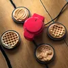 A red mini waffle maker is on a wooden table, surrounded by four finished waffles with different shapes: pumpkin, heart, gingerbread man, and classic round. Produktbild-Vorschau