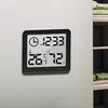 A digital thermometer and hygrometer with a clock function is mounted on a light gray wall, next to a shelf with books. Anteprima dell’immagine prodotto
