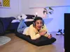 A young man is playing a video game while lying on a floor chair in a bedroom. Náhľad obrázku produktu