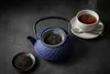 A blue cast iron teapot with a tea strainer filled with loose leaf tea, a small dish of loose leaf tea, and a white cup of tea with a spoon on a saucer, all arranged on a dark textured surface. Podgląd zdjęcia produktu