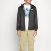 A person wearing a black jacket and khaki pants against a white background. Produktbild-Vorschau
