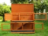 A wooden rabbit hutch with multiple levels and open doors, set on a grassy lawn with trees in the background. Produktbild-Vorschau