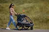 A woman pushing a child trailer on a paved path with grassy hills in the background. Produktbild-Vorschau