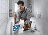 A man is kneeling on the floor of a bathroom, using a power tool to cut a pipe. The bathroom is tiled and has a toilet and shower visible in the background. Podgląd zdjęcia produktu