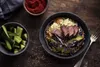 A bowl of ramen with steak, avocado, and red cabbage, served with a side of sliced cucumbers and a small bowl of red chili paste. Produktbild-Vorschau