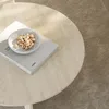 A close-up of a round travertine coffee table with a book and a small bowl of pistachios on it, placed on a carpeted floor. Produktbild-Vorschau