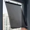 A black mesh roller blind is being attached to a window with suction cups. The blind is partially rolled down, and a hand is visible at the bottom right, holding a suction cup. Voir les images du produit