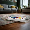 A power strip with multiple outlets is placed on a rug in a living room, with a sofa and window in the background. Produktbild-Vorschau