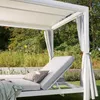 A white outdoor lounge chair with gray cushions and curtains, set in a garden with lush greenery and yellow flowers. Produktbild-Vorschau