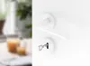 A white door handle with a key in the lock, set against a white background. The left side of the image shows a blurred interior scene with plants and candles. Produktbild-Vorschau