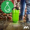 A person in jeans and rubber boots is holding a green submersible pump in a flooded area with a stone wall in the background. A green circle with a water droplet icon is in the upper left corner. Podgląd zdjęcia produktu