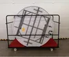 A round folding table is stored vertically on a black metal cart with a red base. The cart has wheels and is positioned in front of a white garage-style door. Produktbild-Vorschau