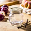 A small glass jar with a metal lid sits on a wooden surface, surrounded by plums and vanilla beans. Produktbild-Vorschau
