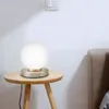 A modern globe-shaped table lamp with a silver base sits on a small wooden side table next to a bed. Produktbild-Vorschau