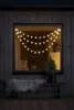 A string of star-shaped LED lights hangs across a window of a wooden cabin, with a bench and a small tree outside. Produktbild-Vorschau