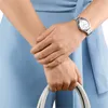 A close-up shot of a woman's arms and hands, one wearing a silver watch, holding a white handbag. Náhled obrázku produktu
