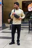 A man stands outdoors wearing a beige sweater, black pants, and black sneakers, with a black fanny pack across his chest. Voir les images du produit