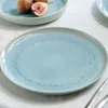 A close-up view of a light blue plate with a textured pattern, set on a white tablecloth. Another plate with figs is visible in the background. Náhľad obrázku produktu
