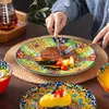 A person is cutting a steak on a colorful, patterned plate, with other dishes and a drink in the background. Náhled obrázku produktu