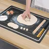 A person is using a star-shaped cookie cutter on dough placed on a black silicone baking mat with measurement markings. Other baking ingredients and tools like eggs, flour, and a rolling pin are also visible on the mat and the wooden table. Voir les images du produit