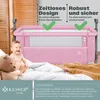 A baby is sitting in a pink crib next to a teddy bear, with blankets and pillows in the foreground. Produktbild-Vorschau