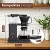 A coffee maker is shown on a wooden countertop with a brick wall and shelves in the background. Produktbild-Vorschau