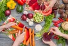 A top-down view of a collection of fresh vegetables and hands preparing them on a wooden table. Produktbild-Vorschau