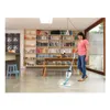 A woman is using a steam mop to clean a floor in a bright room with large bookshelves. Voir les images du produit