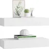 Two white floating shelves are displayed against a white background. The top shelf holds a framed picture of leaves, a small potted cactus, and a set of reed diffusers. The bottom shelf displays two books with a decorative white vase on top. Produktbild-Vorschau