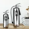 Two polished metal fire extinguishers are displayed on a wooden surface in front of a white background. A vase with cotton branches is on the right. Produktbild-Vorschau
