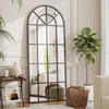 A full-length arched mirror with a black metal frame is shown in a well-lit room, reflecting a sofa and other decor. Plants and a small side table with a vase and magazine are also visible. Produktbild-Vorschau