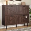 A dark wood cabinet with a barn door design, featuring a cat peeking out of one of the compartments. The cabinet is placed in a room with a lamp, books, and a handbag on top. Produktbild-Vorschau