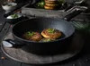 A frying pan with food being cooked, set on a wooden surface with various ingredients and bowls in the background. Produktbild-Vorschau