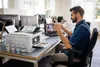 A man is sitting at a desk in an office, interacting with a printer and a laptop during a video call. Náhled obrázku produktu