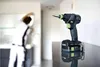 A cordless drill is placed on a reflective surface in the foreground, with a person blurred in the background working on a wall. Voir les images du produit