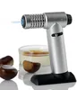 A silver and black culinary torch with a blue flame, positioned next to a crème brûlée and chestnuts. Produktbild-Vorschau