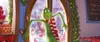 The Grinch is peeking through a doorway decorated with Christmas lights and garland. There are cakes and other festive items visible in the background. Náhľad obrázku produktu