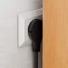 A black electrical plug is inserted into a white wall socket, partially obscured by a wooden surface. Produktbild-Vorschau