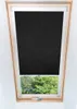 A black roller blind is installed in a wooden-framed attic window, with green trees visible outside. Náhled obrázku produktu