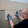 A person is tiling a wall with large beige tiles. Anteprima dell’immagine prodotto