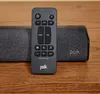 A close-up shot of a Polk Audio soundbar and its remote control, placed on a wooden surface. Produktbild-Vorschau